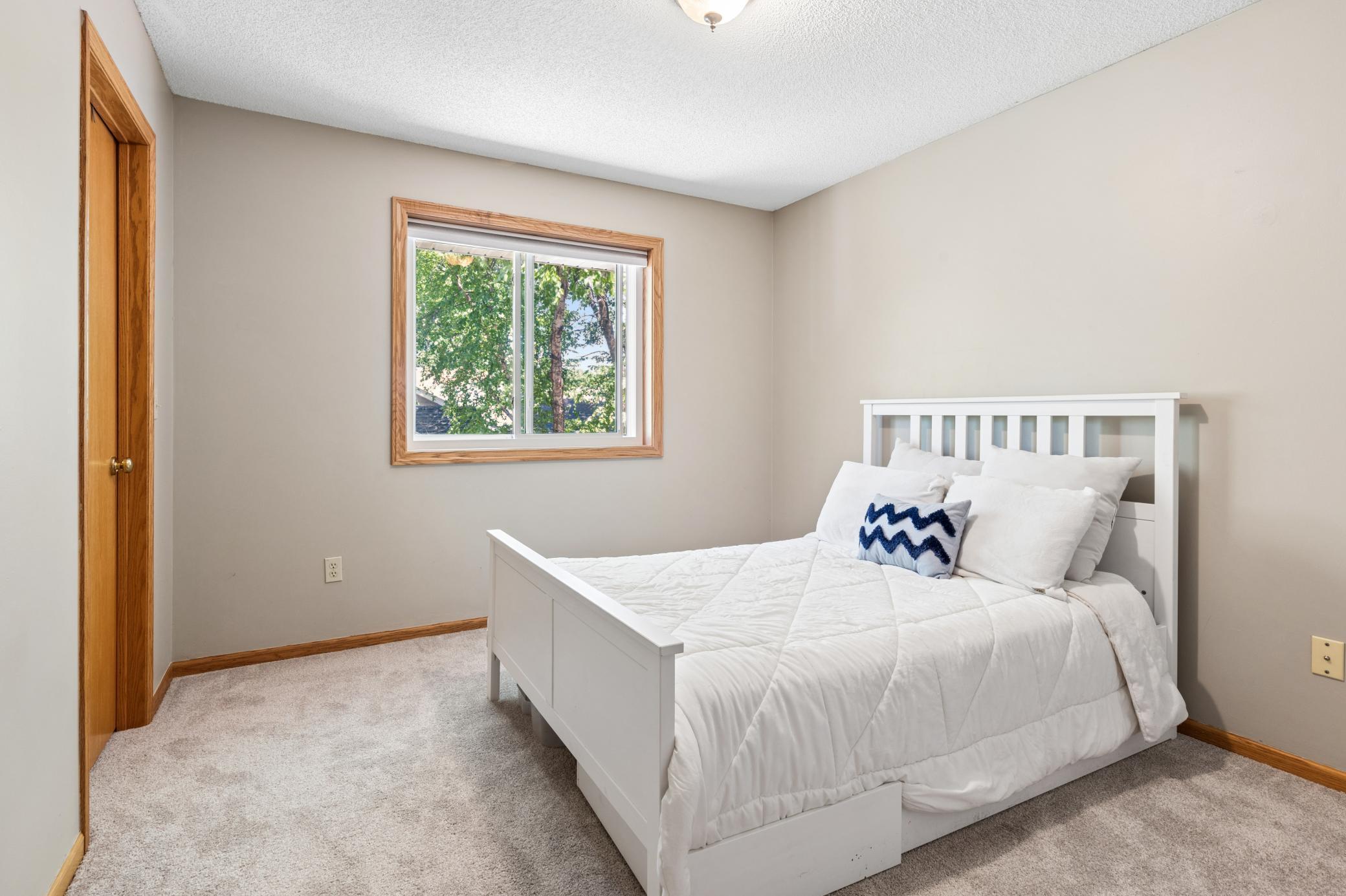 This upper floor second bedroom is quiet, calm, and ready for whoever comes next. White frame bed. Soft carpet. A tree-filled window that keeps the room feeling connected to the outdoors.