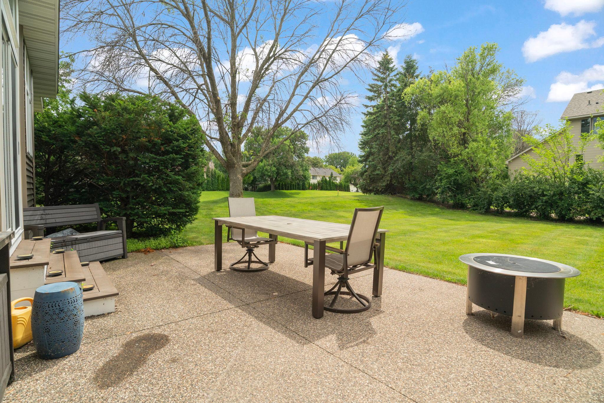 The patio opens to a wide green yard with mature trees lining every edge. A fire pit ready for tonight. The world past the fence is quiet. This is the view you'll come home for.