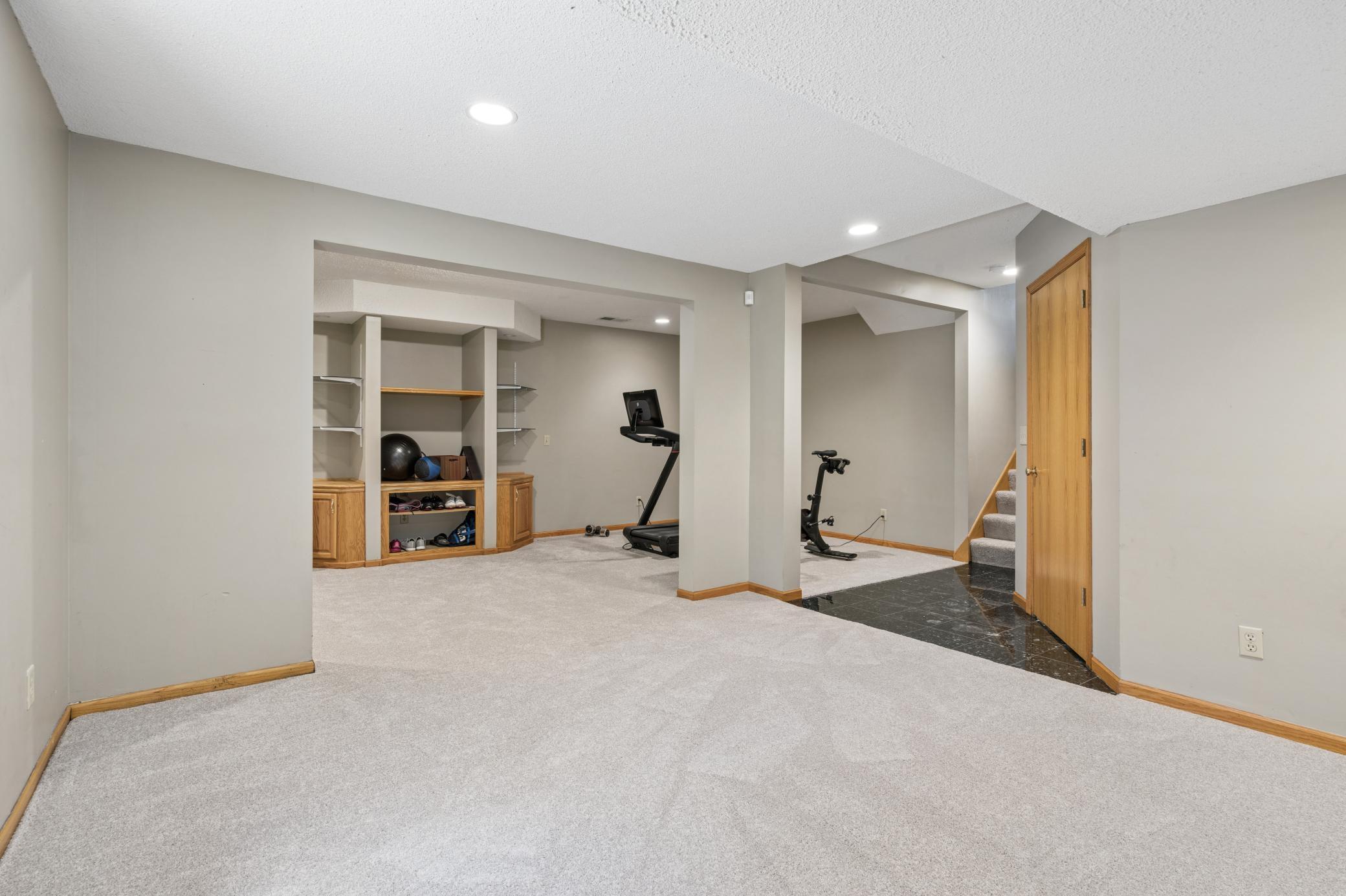 The full picture of what this lower level offers. A wide open family room up front, a dedicated workout zone tucked behind the columns, built-in storage, and stairs back up to the rest of your life.