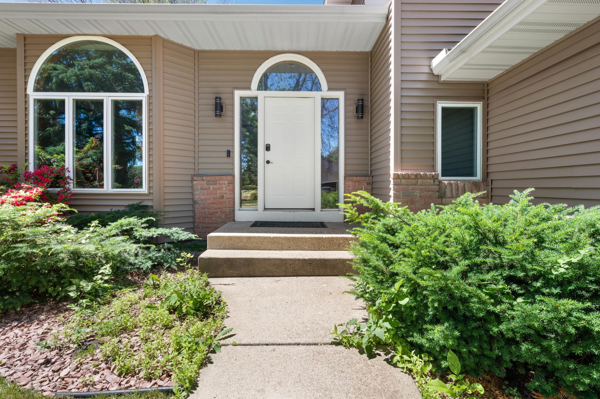 Two arched windows flank the front door like bookends. Red blooms in the landscaping. Brick at the base. New siding above. The entry says something before you ring the bell.