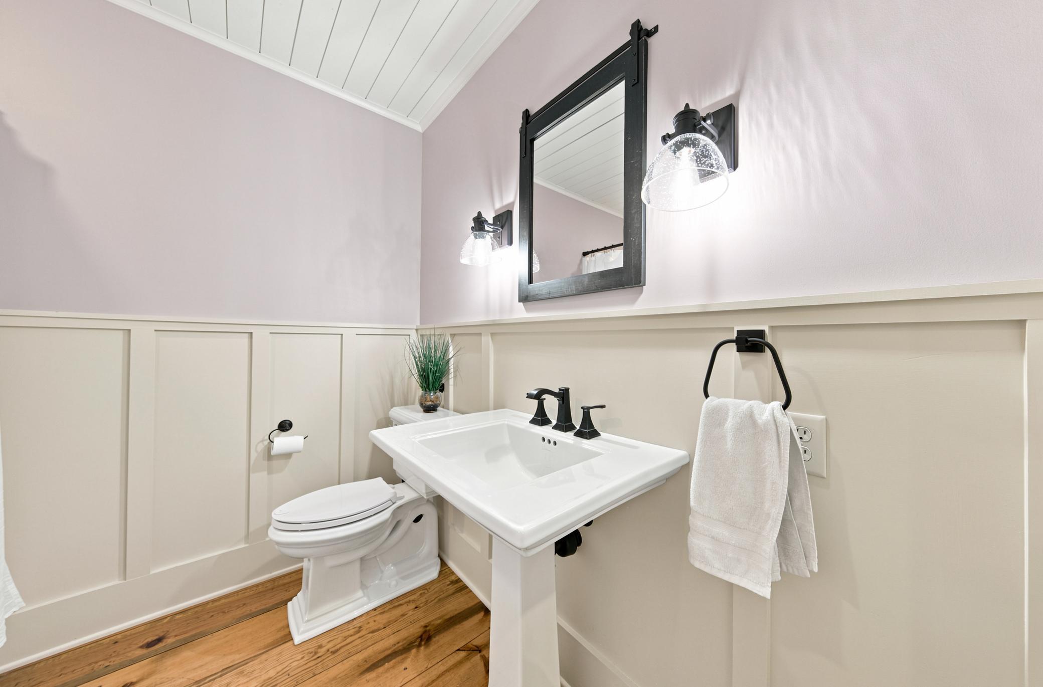 This charming farmhouse-style bathroom on main floor features elegant wainscoting, wide-plank wood floors, a classic pedestal sink, and custom modern light fixtures.