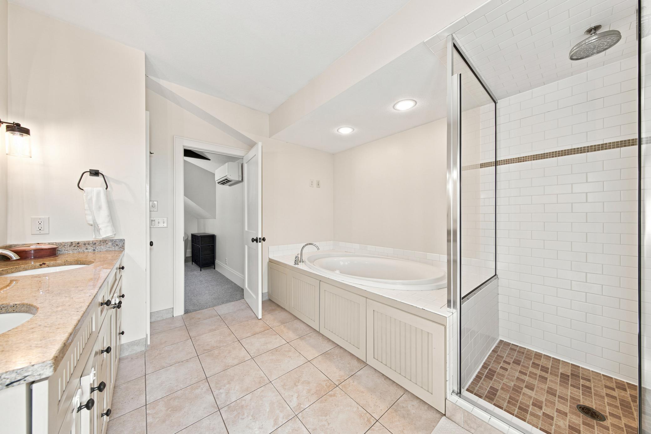 This luxurious bathroom serves both the main bedroom, second bedroom and the adjoining bonus space, featuring a double vanity with stone countertops, modern oval mirrors, a deep soaking tub, and a separate glass-enclosed tile shower.