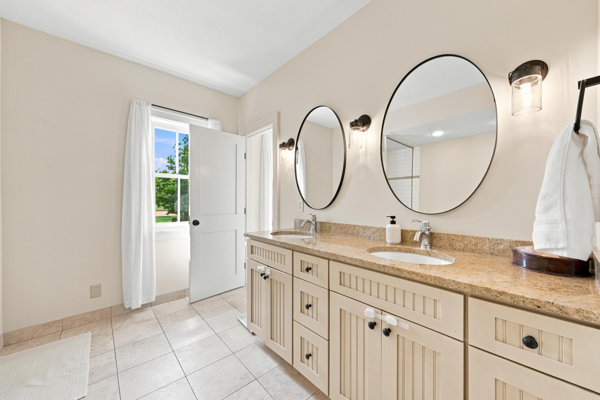 This luxurious bathroom serves both the main bedroom, second bedroom and the adjoining bonus space, featuring a double vanity with stone countertops, modern oval mirrors, a deep soaking tub, and a separate glass-enclosed tile shower.