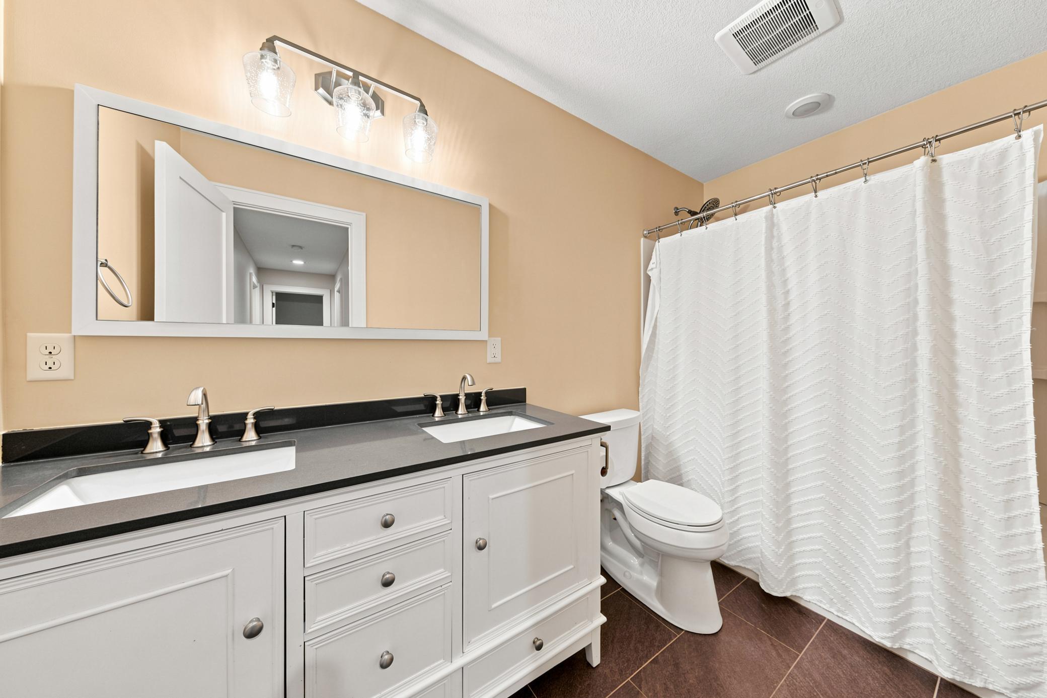 This convenient second main-floor bathroom features a stylish double vanity with dark countertops, a large framed mirror with modern vanity lighting, and durable dark tile flooring.