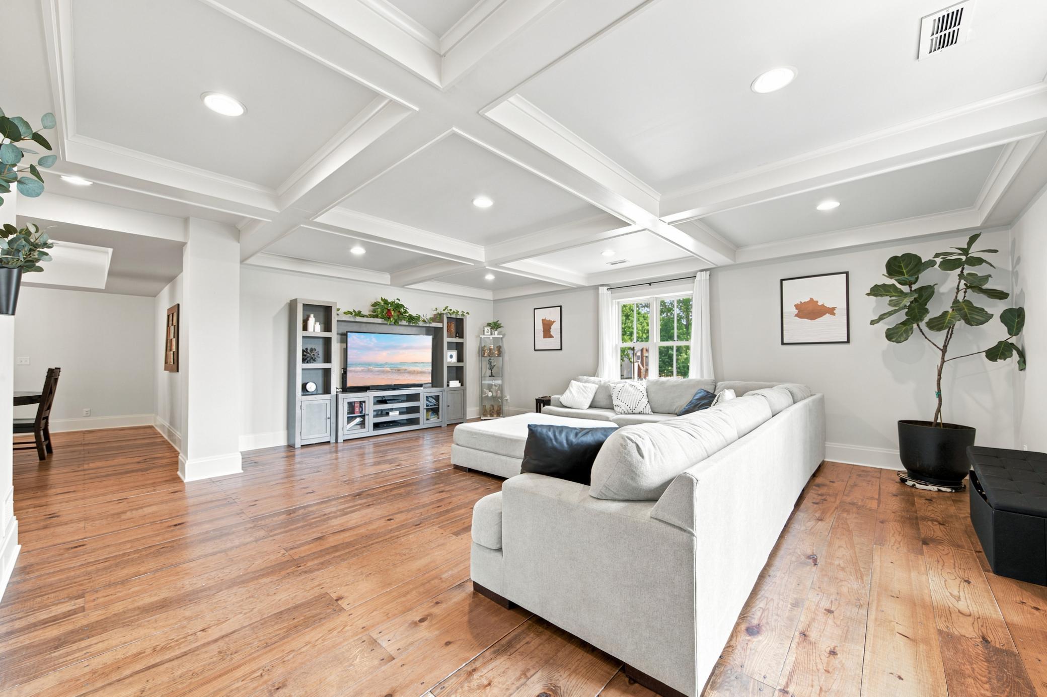 Spacious living room featuring stunning custom coffered ceilings, rich wide-plank hardwood floors, and large windows framing lush outdoor views.