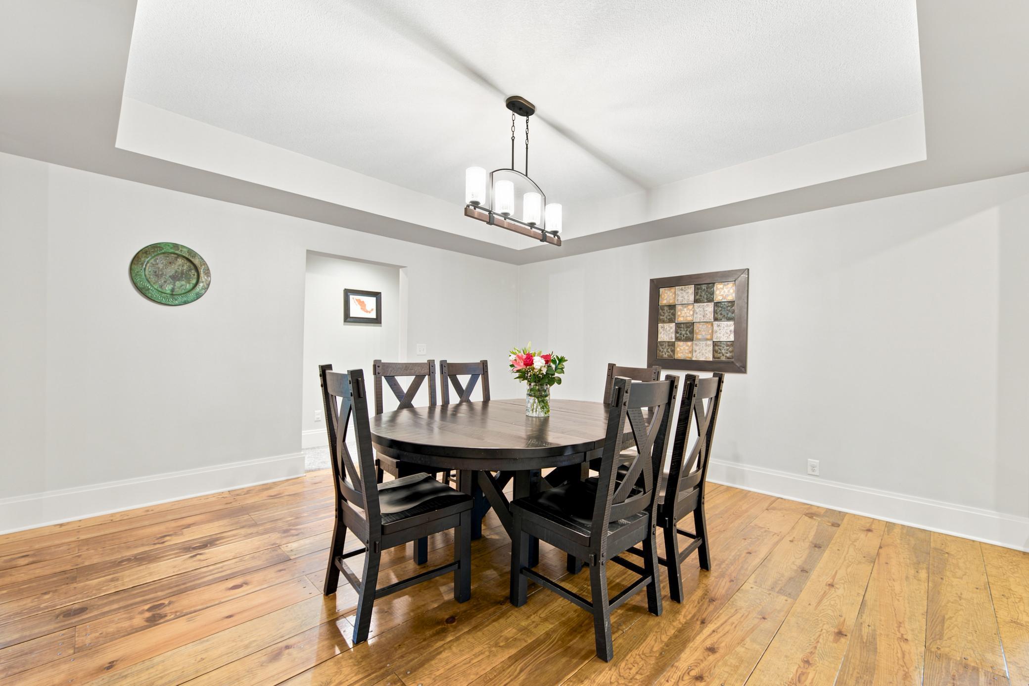 An elegant, open-concept dining area showcases rich wide-plank wood floors, a stylish tray ceiling, an exposed brick detail, and an effortless transition into a modern chef's kitchen.