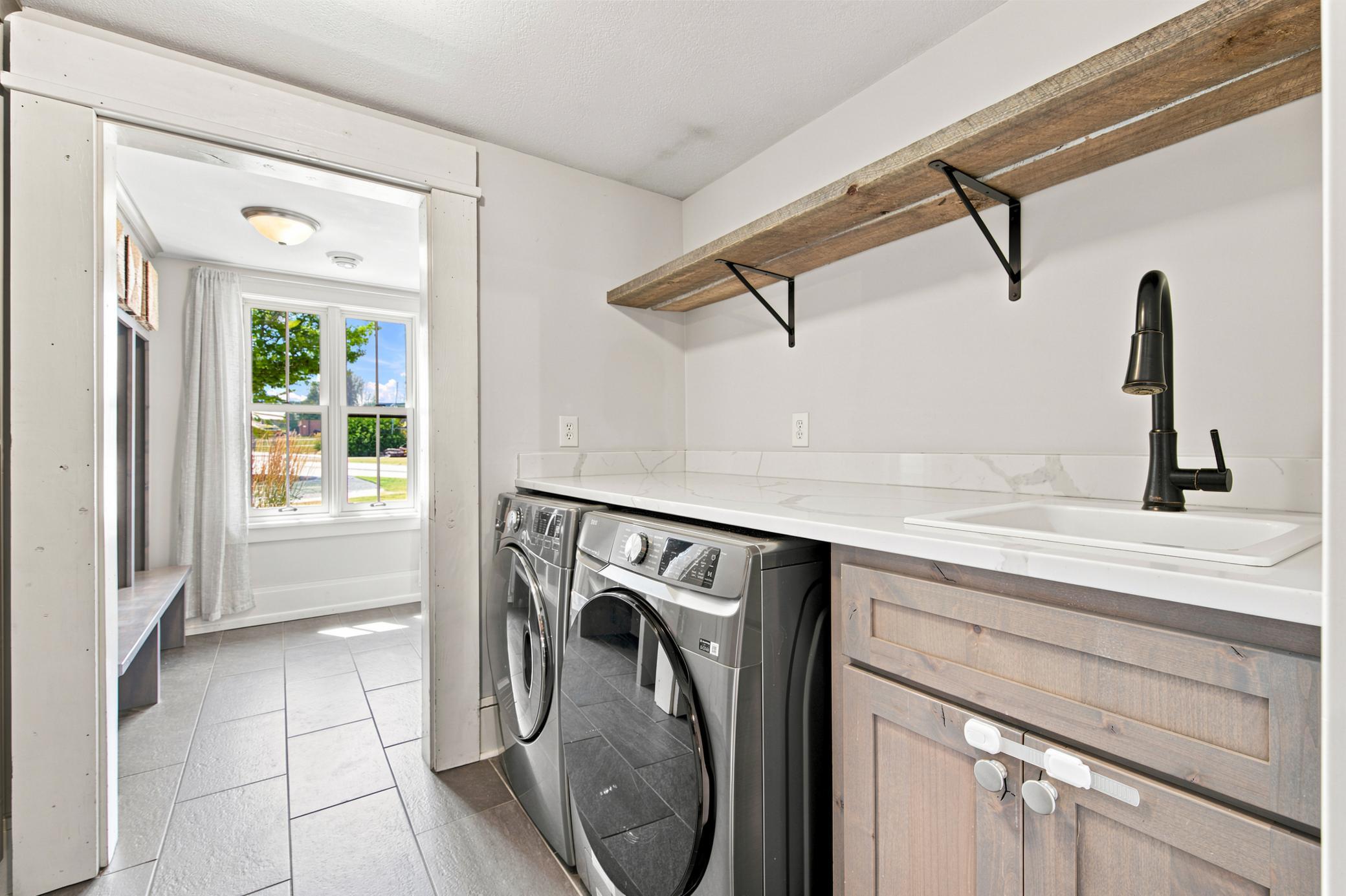 This highly functional main-floor laundry room features a premium washer and dryer set, a utility sink with quartz countertops, durable tile flooring, and an abundance of floor-to-ceiling storage cabinets.