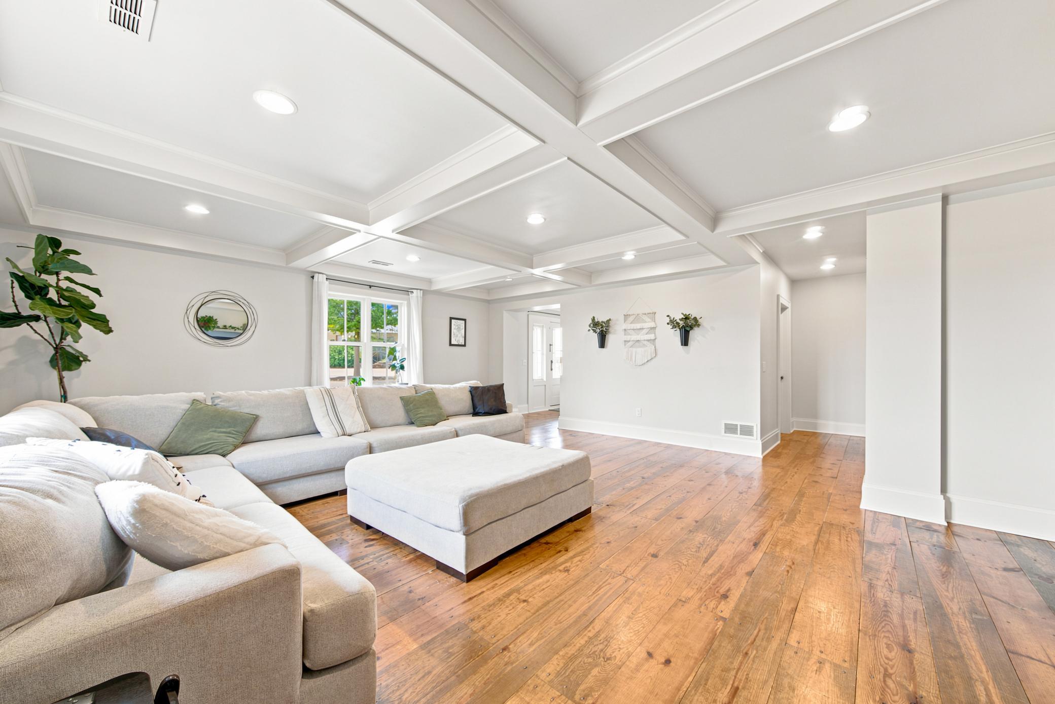 Spacious living room featuring stunning custom coffered ceilings, rich wide-plank hardwood floors, and large windows framing lush outdoor views.
