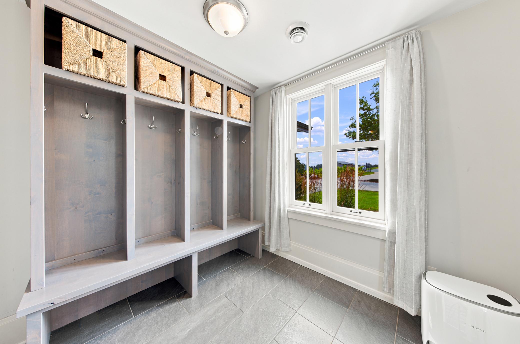A bright, functional mudroom features expansive custom built-in lockers with coat hooks, upper basket storage, a built-in bench, and a large window offering ample natural light over durable tile floors.