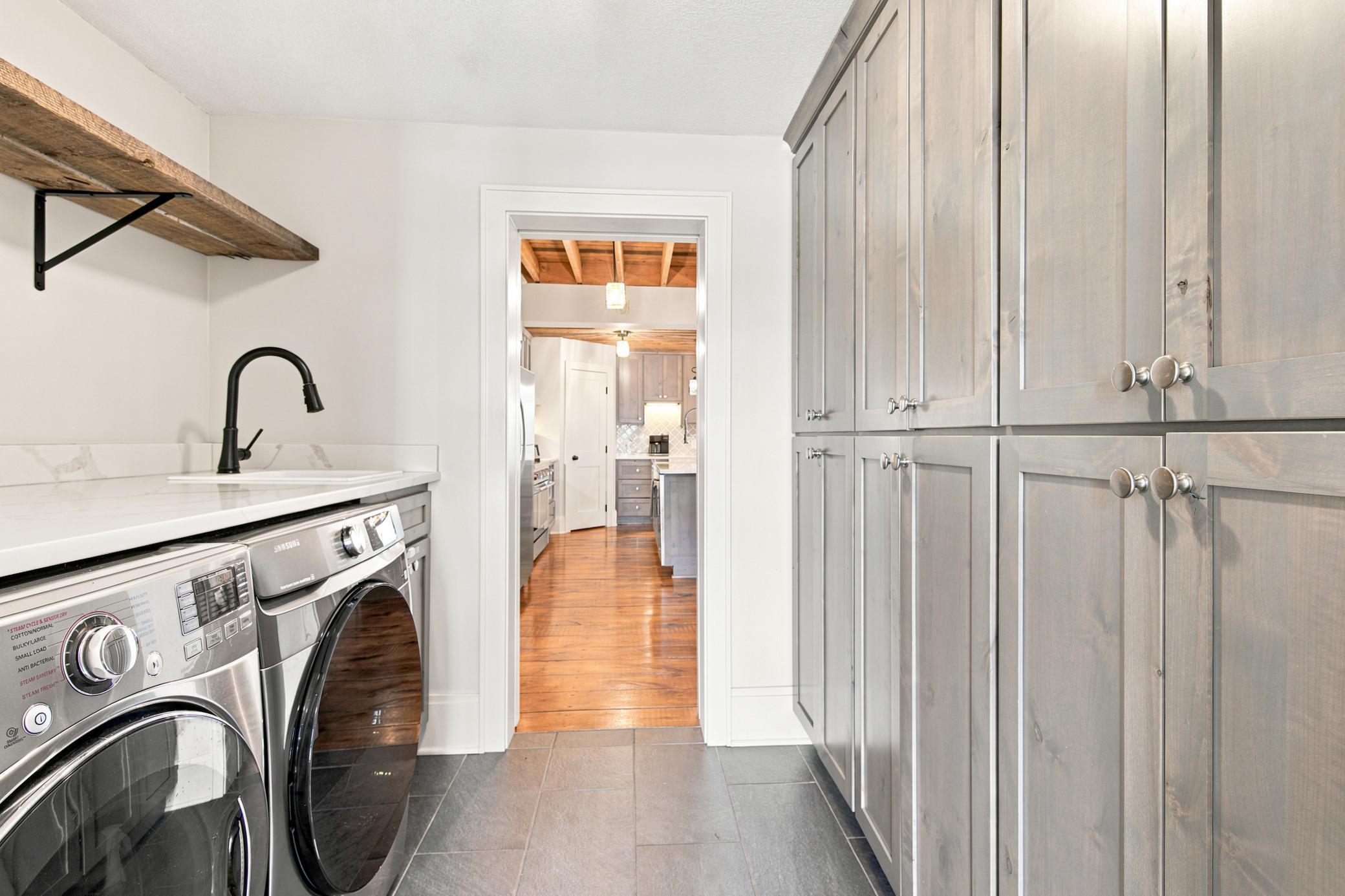 This highly functional main-floor laundry room features a premium washer and dryer set, a utility sink with quartz countertops, durable tile flooring, and an abundance of floor-to-ceiling storage cabinets.