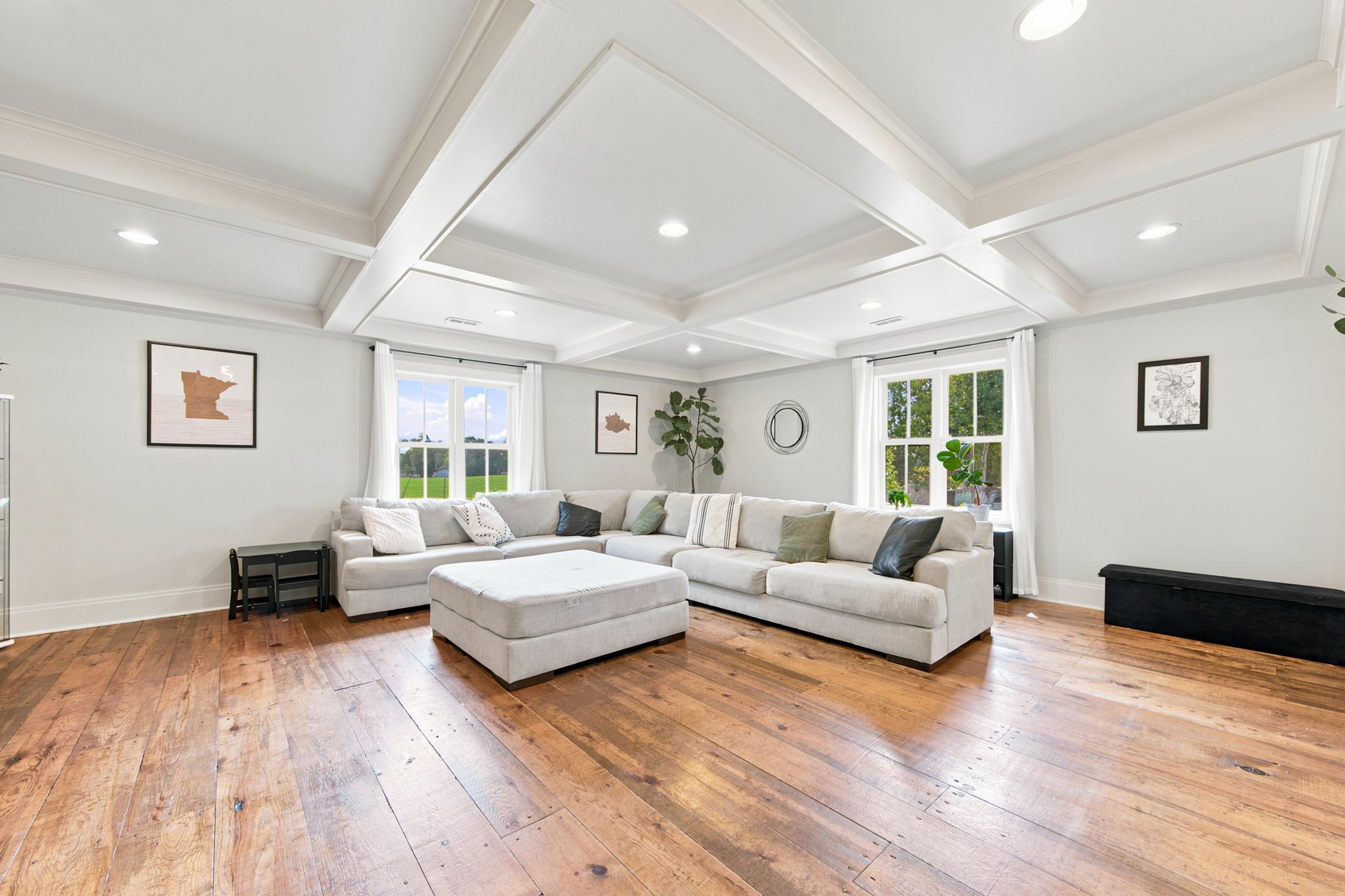 Spacious living room featuring stunning custom coffered ceilings, rich wide-plank hardwood floors, and large windows framing lush outdoor views.