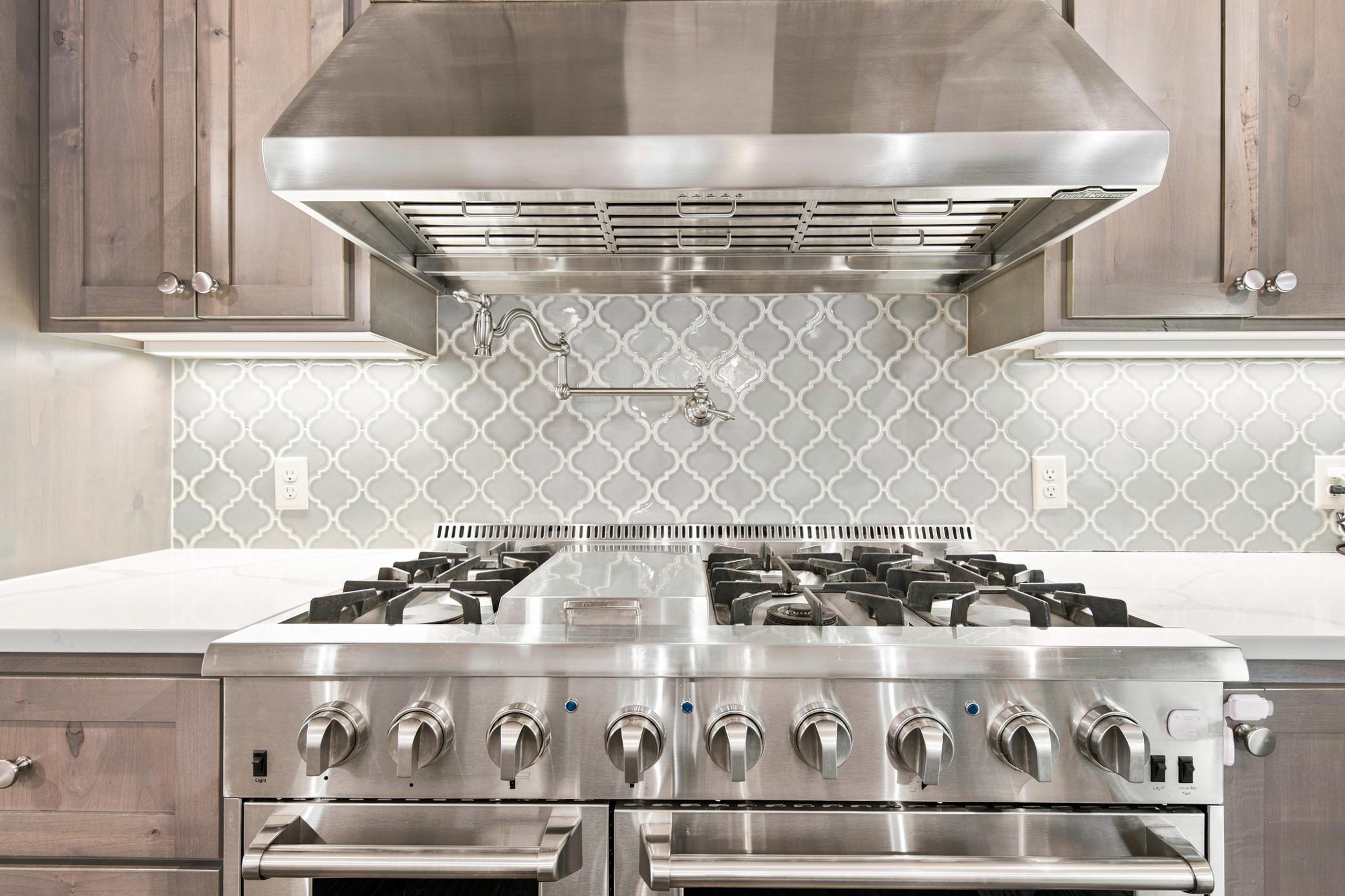 A commercial-grade stainless steel gas range is perfectly accented by a stylish arabesque tile backsplash, a convenient pot filler, and a professional-grade vent hood flanked by custom cabinetry.