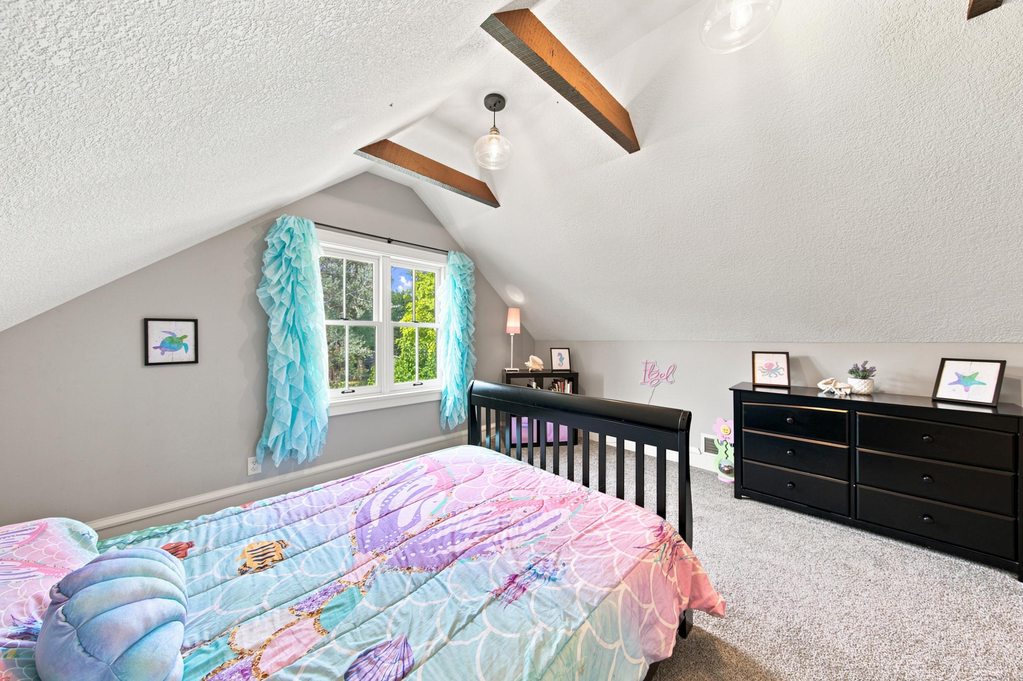 This charming second bedroom connects directly to the flexible bonus space as well and features a dramatic vaulted ceiling with rustic wood beams, plush carpeting, and a bright window with scenic views.