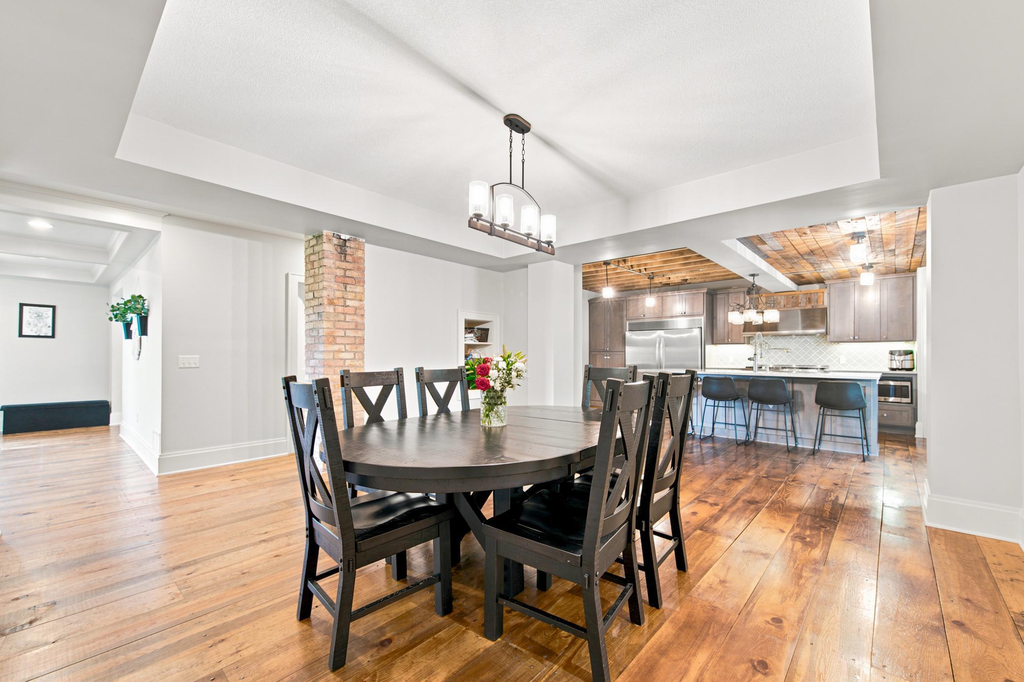 An elegant, open-concept dining area showcases rich wide-plank wood floors, a stylish tray ceiling, an exposed brick detail, and an effortless transition into a modern chef's kitchen.