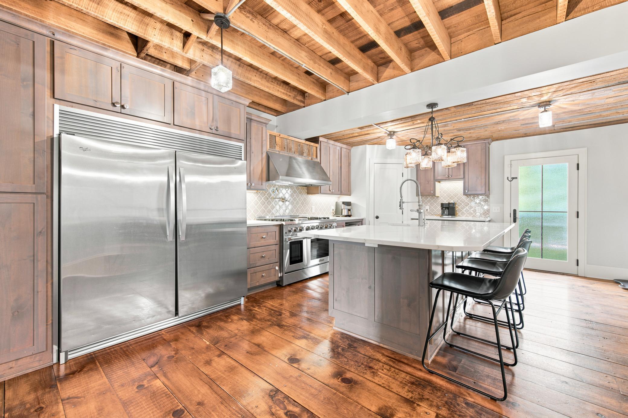 This gourmet kitchen boasts high-end stainless steel appliances, premium quartz countertops with a farmhouse sink, custom cabinetry, and dramatic exposed wood-beam ceilings.