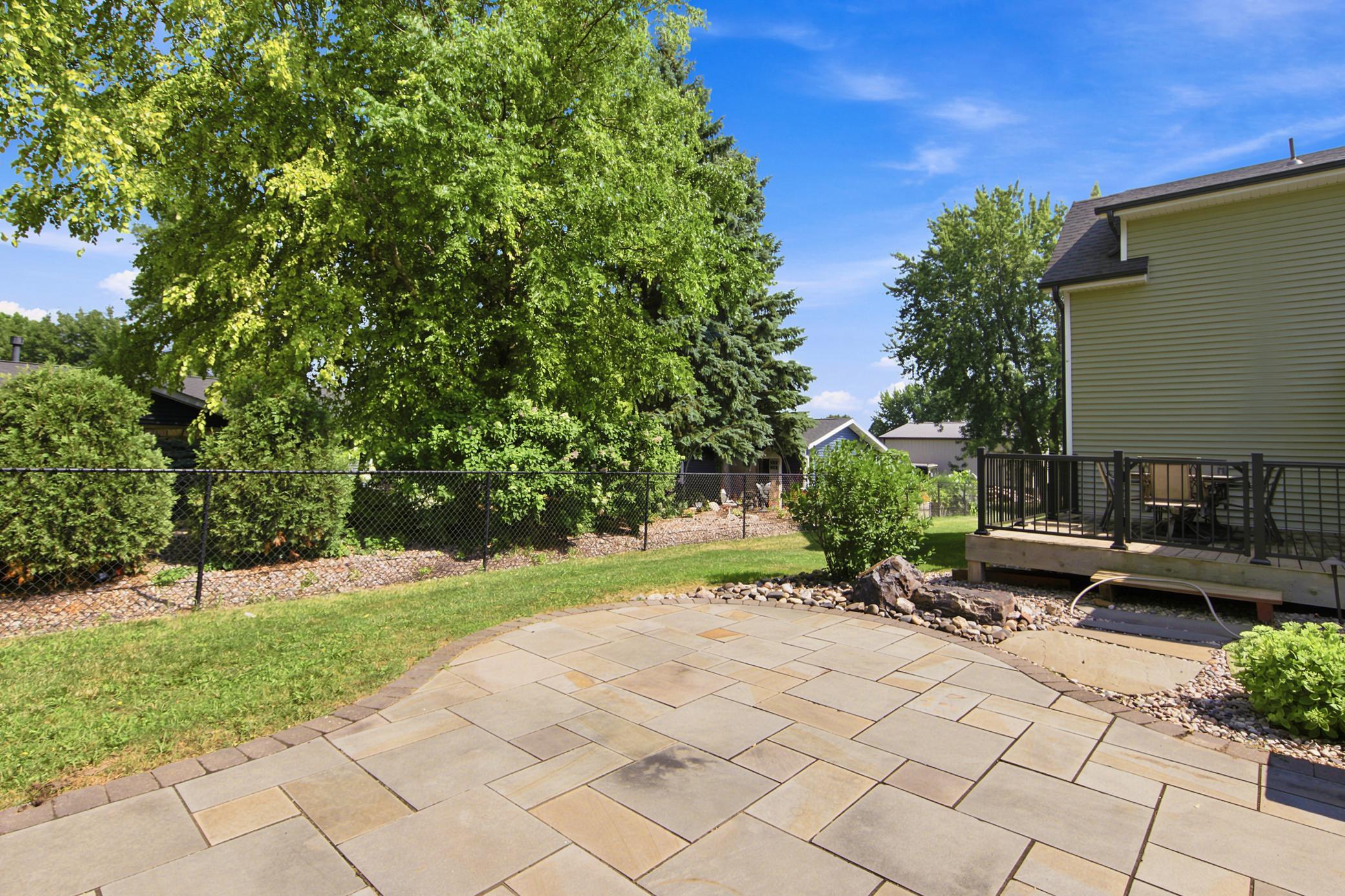 The exceptional outdoor living space features an elevated wood deck overlooking a beautifully landscape, complete with a custom stone patio, a low retaining wall, and mature privacy trees.