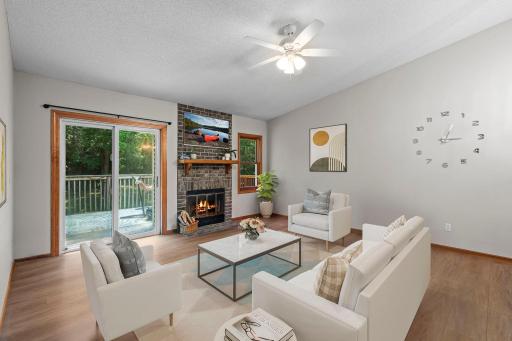 The spacious living room showcases new luxury plank flooring (2023), a new ceiling fan, a cozy wood-burning fireplace with brick surround, and access to an 11x11 deck. *This Image Is Virtually Staged*
