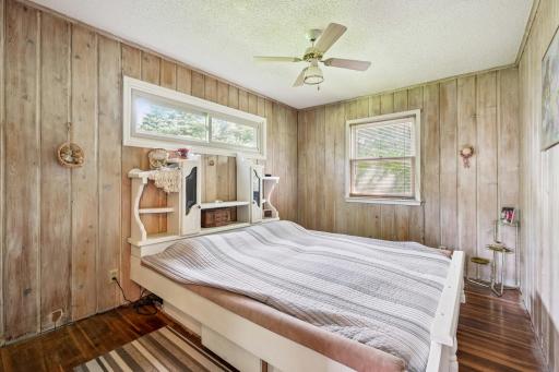 Second bedroom with wood panel walls, ceiling fan, and multiple windows overlooking the yard.
