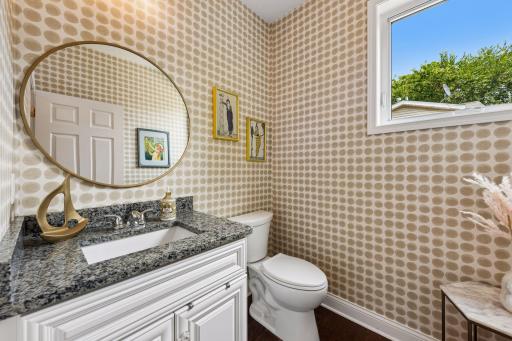 This charming powder room features granite countertops, elegant cabinetry, designer wallpaper, and a bright window that fills the space with natural light and character.