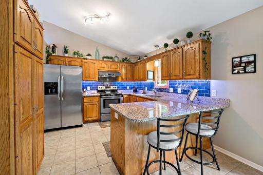 The spacious kitchen features an abundance of cabinetry, granite countertops, tile floors, a striking blue tile backsplash, stainless steel appliances, and ample counter space.