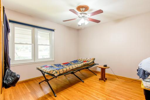 Second main-level bedroom with hardwood flooring, a ceiling fan, and large windows overlooking the front yard.