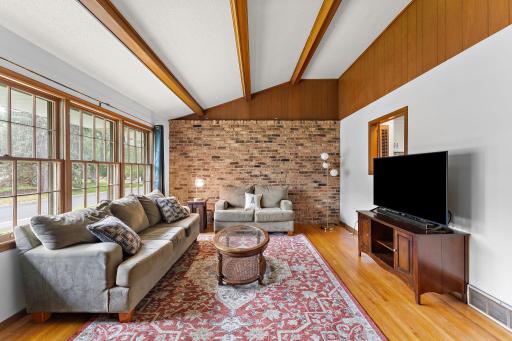 The front living room of the home with the front yard and street visible through the windows on the left. The front room features vaulted ceilings with exposed wood beams and a brick accent wall. This is the main/third floor.