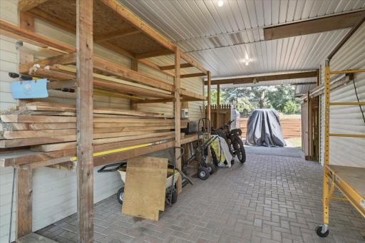 Covered breezeway-style carport provides protected parking and additional storage, complete with durable paver flooring, built-in lumber racks, pull down attic ,and convenient access between the heated garage, workshop, studio, and backyard.