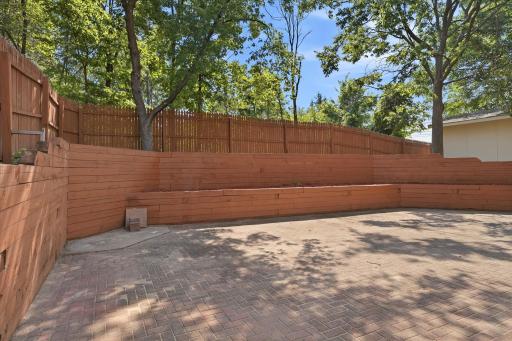 Private courtyard-style patio features an expansive paver surface surrounded by attractive tiered retaining walls and mature trees, creating a secluded outdoor space ideal for entertaining, relaxing, or enjoying a quiet evening outdoors.