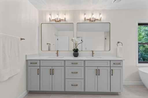 Luxurious primary bathroom featuring a dual vanity with custom gray cabinetry, quartz countertops, gold fixtures, and designer lighting above dual mirrors.