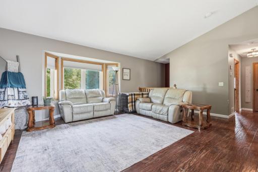 This spacious living room offers vaulted ceilings, hardwood flooring, and a bay window overlooking mature trees for a bright, private feel. Comfortable seating and an easy flow to the hallway make it a natural gathering spot.