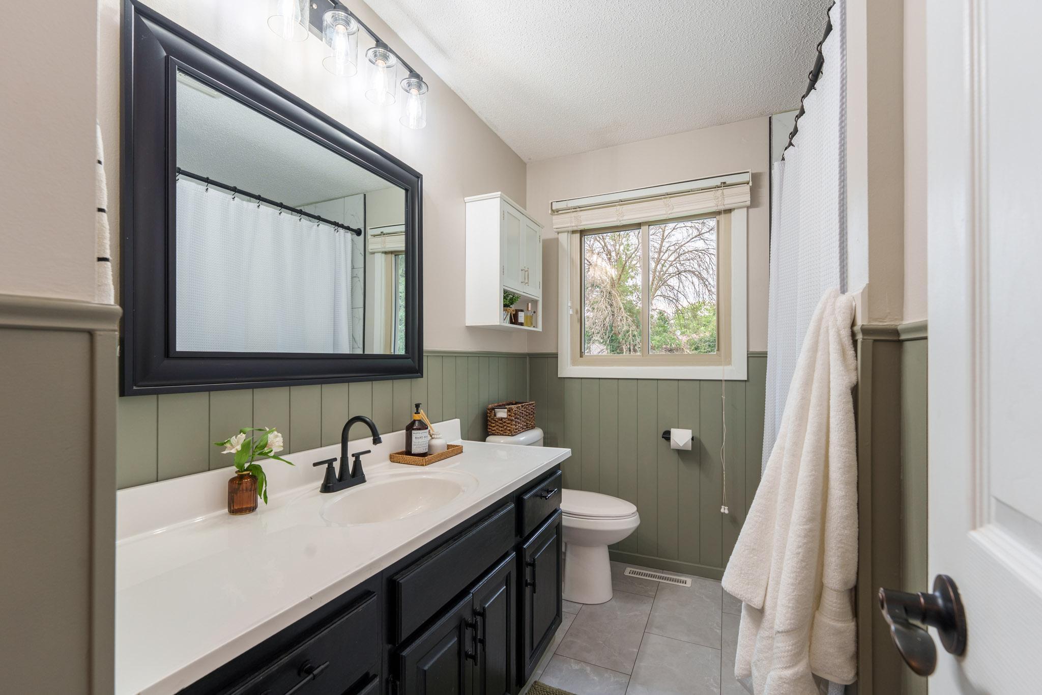 Beautifully remodeled bathroom, completed in 2025, featuring a tiled shower, wainscoting detail, new flooring, updated vanity, & fixtures.