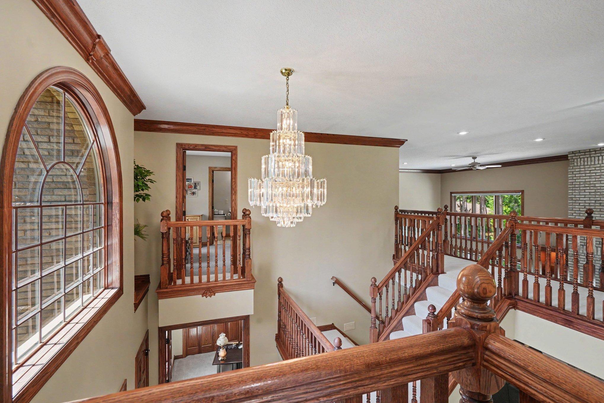 An elegant upper-level overlook showcases rich wood railings and a striking chandelier on a convenient lowing system cleaning and bulb changes. The open staircase adds drama while offering a beautiful view into the main entry below.