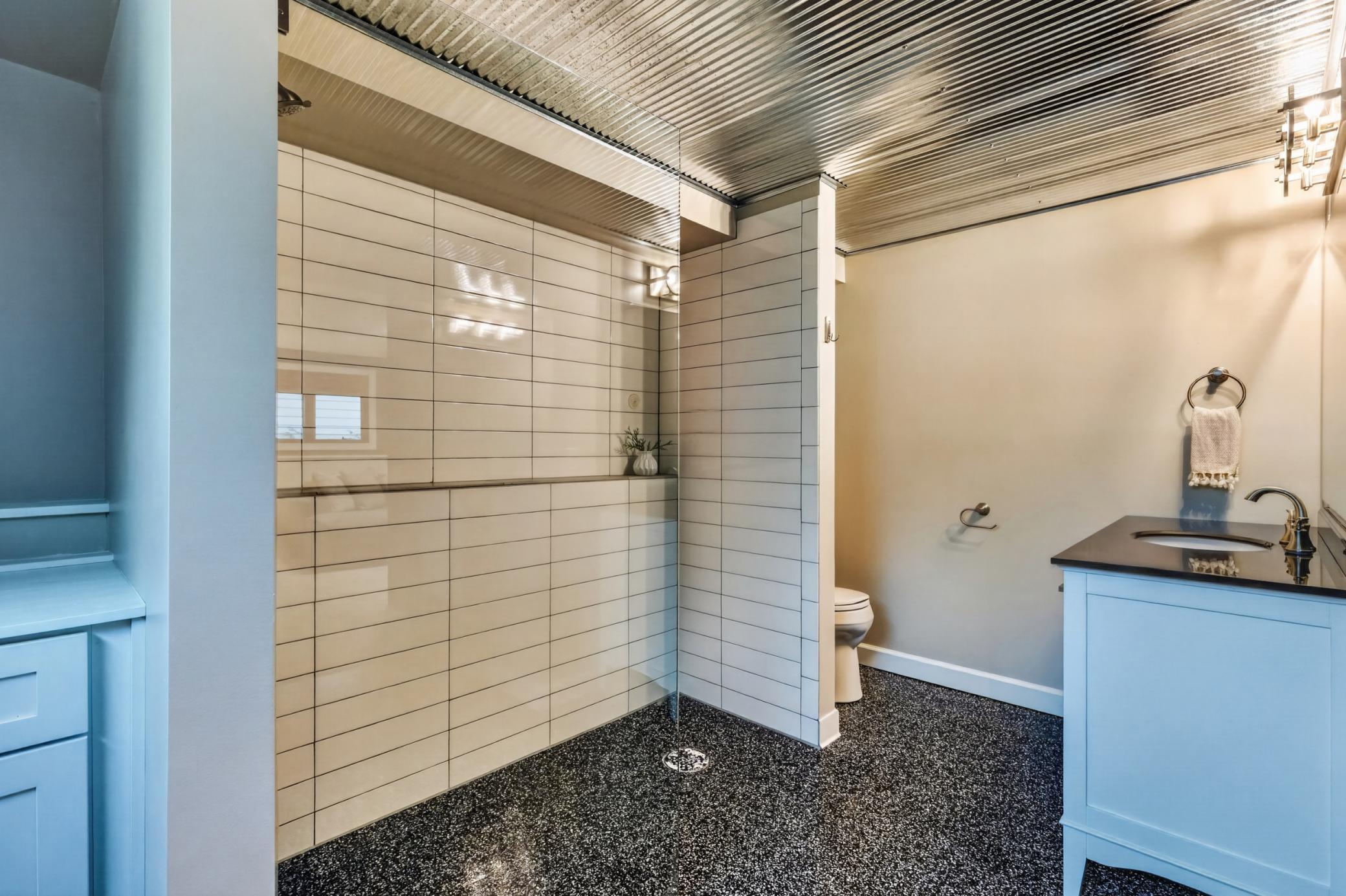 Lower 3/4 bathroom features an enameled vanity cabinet and additional storage cabinet w/ quartz tops. Oversized walk-in shower features zero entry, subway tile surround, shampoo niche & glass panel. Corrugated tin ceiling & epoxy coated flooring