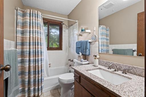 The full bathroom on the upper level features a granite-topped vanity, generously sized mirror, tiled tub and shower surround, and a window that brings in natural sunlight.