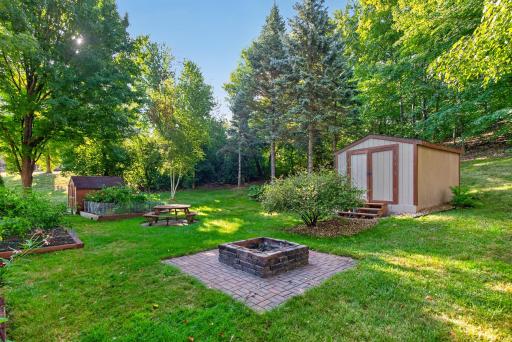 The wooded yard offers multiple ways to enjoy the outdoors, with a brick-paver fire pit, picnic area, garden beds, and two storage sheds surrounded by mature trees.