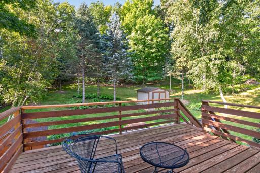 The elevated deck overlooks a beautifully wooded backyard, where mature trees, open lawn, and two storage sheds create a private and peaceful outdoor setting.