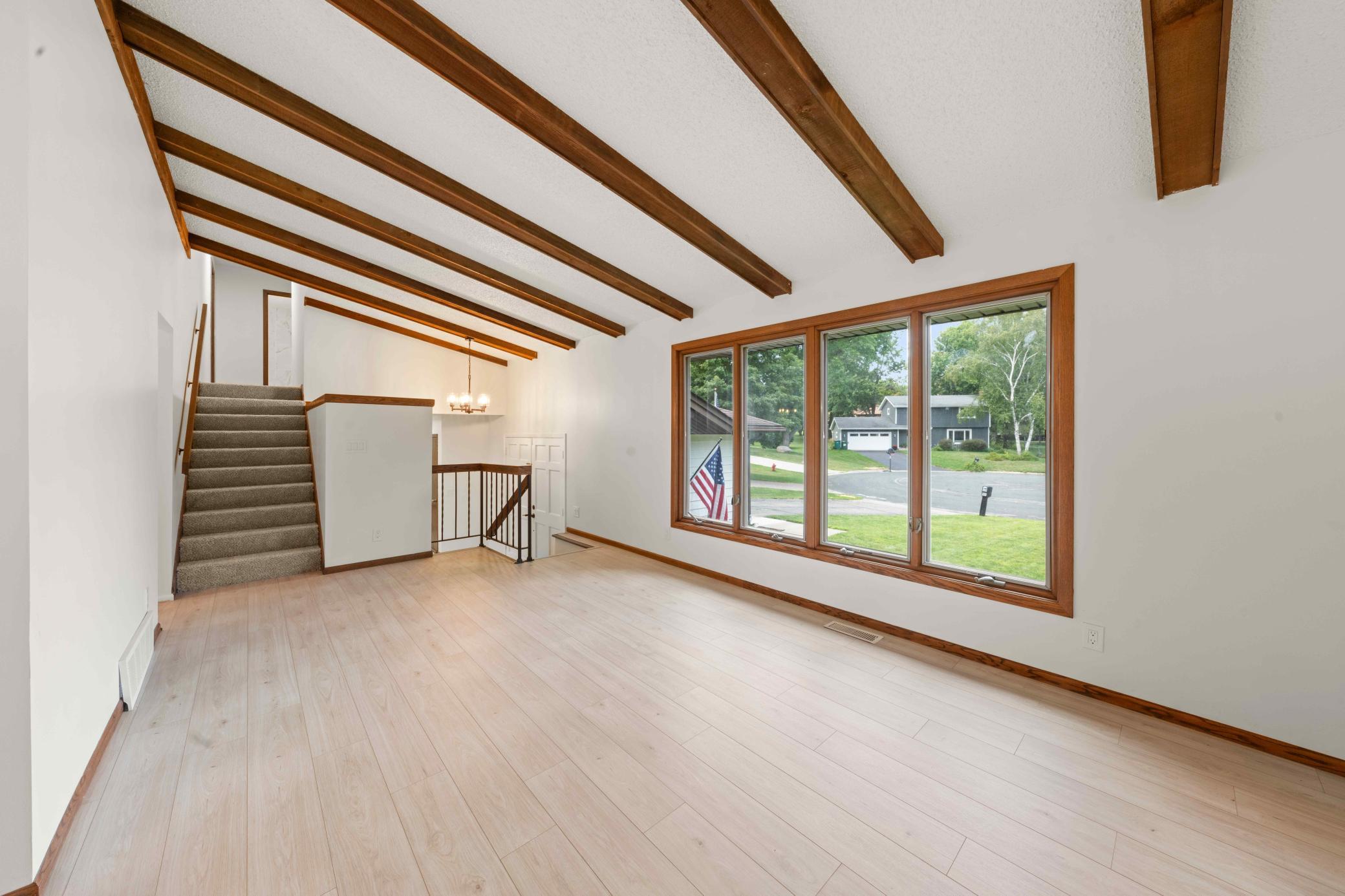 Spacious living room with vaulted exposed-beam ceiling and a wall of windows bringing in abundant natural light. Staircase to upper level
