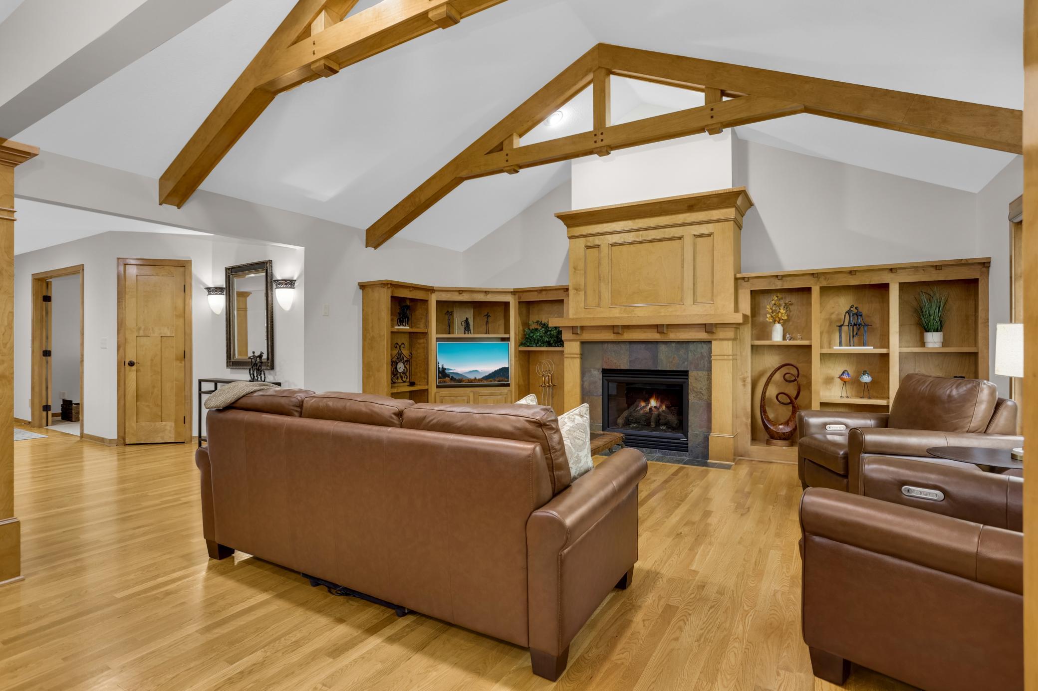 Inviting living room features beautiful maple-built-ins, a glowing fireplace, and stunning vaulted beamed ceilings that create a warm and impressive gathering space