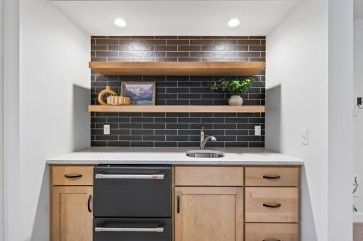 This stylish wet bar boasts striking dark tile and floating shelves, plus a sink and fridge; the perfect spot to grab a drink after some friendly competition on the sport court.