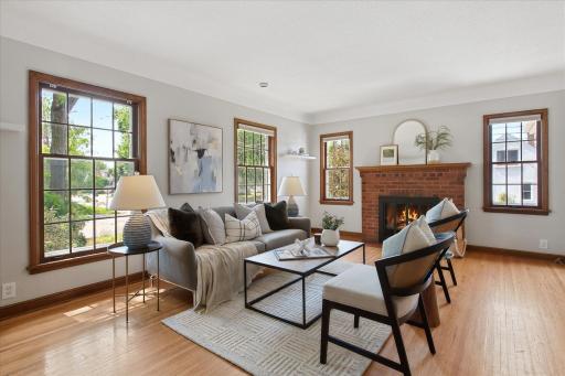 The spacious living room centers around a cozy wood-burning fireplace with a classic brick surround, while multiple windows provide natural light and leafy neighborhood views.