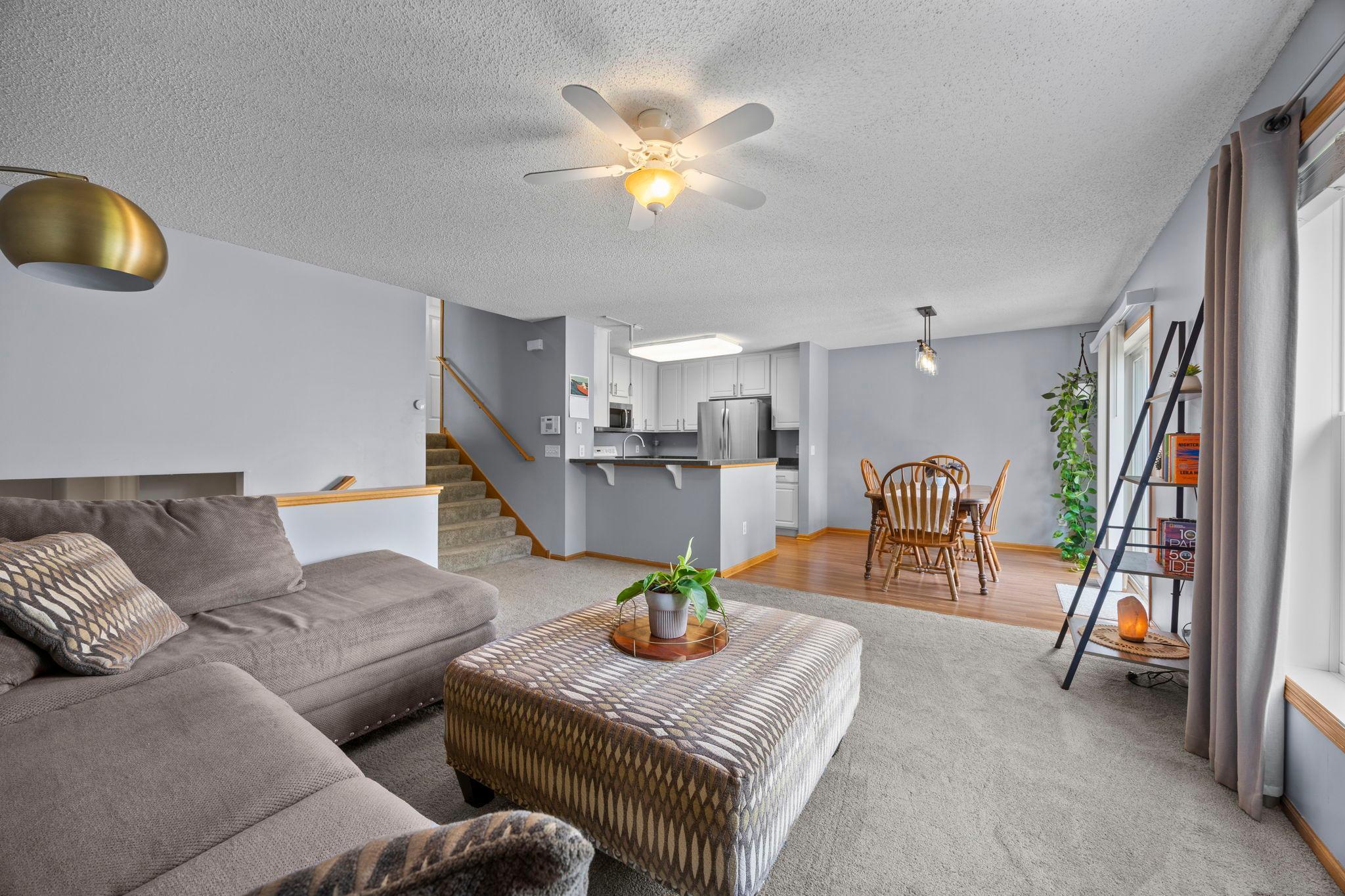 The living room features soft carpeting and a large window, flowing naturally into the kitchen and dining area.