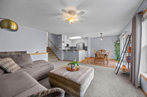 The living room features soft carpeting and a large window, flowing naturally into the kitchen and dining area.