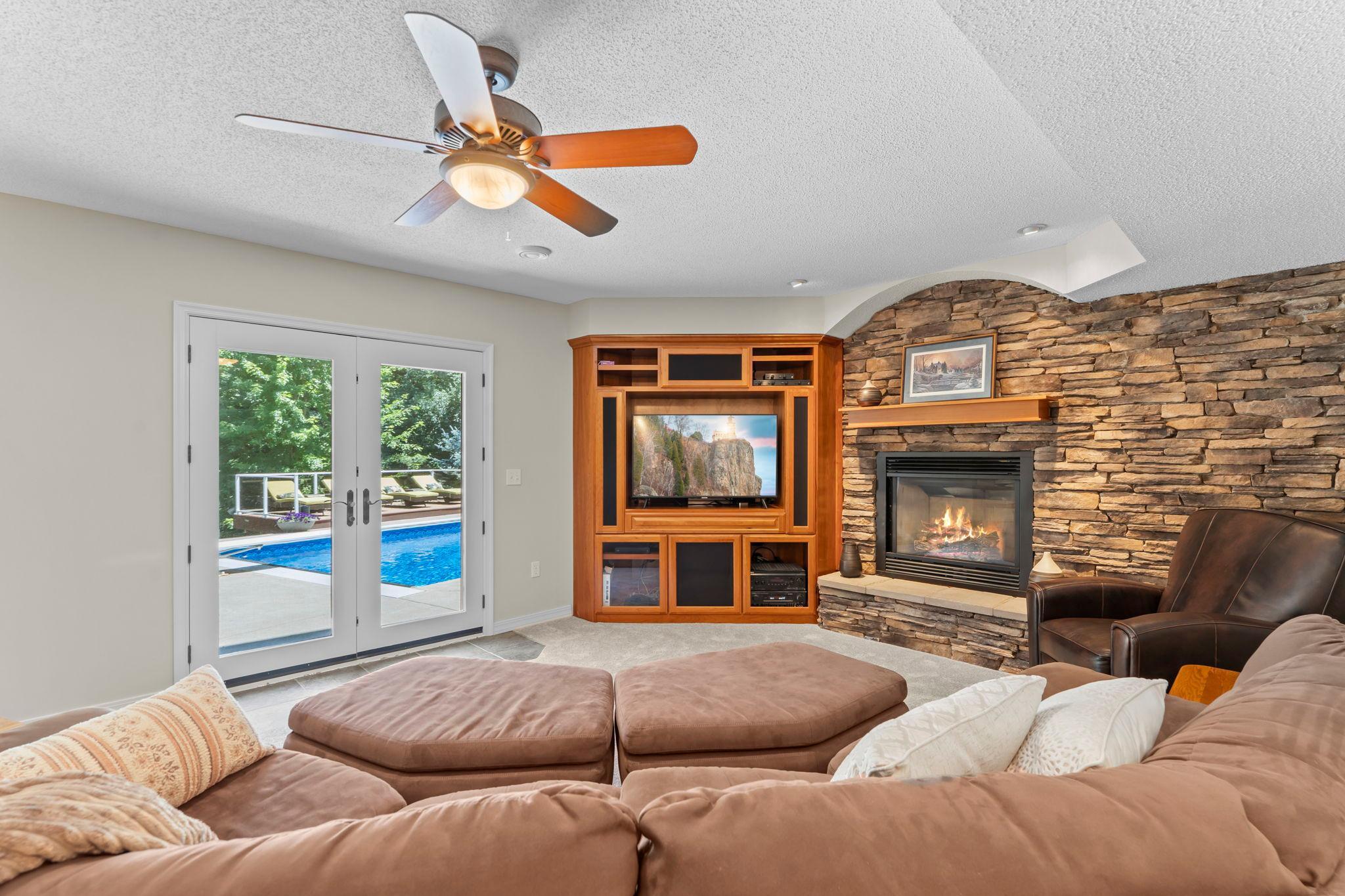 The lower-level family room provides a comfortable gathering space with a built-in entertainment center, gas fireplace with stone surround, ceiling fan, and double-door walkout access to the backyard pool deck.