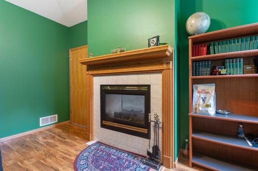 Inviting main-floor office featuring plank flooring, a stunning double-sided glass fireplace with tiled surround, and large windows framing beautiful backyard views—an inviting space to work, relax, or enjoy the scenery.