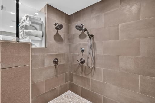 Custom walk-in shower features modern tile, dual shower heads with hand shower, decorative stone mosaic floors, built-in bench, shampoo niches and glass enclosure