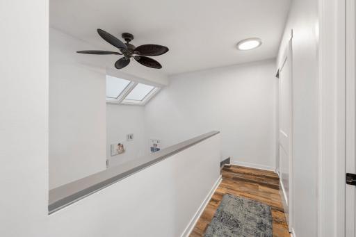 Bright upper hallway featuring continued wood-look flooring, a ceiling fan, and natural light from the skylights.