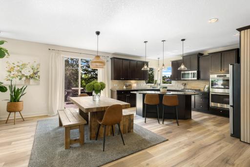 Open and inviting kitchen and dining area featuring updated flooring, dark cabinetry, stainless steel appliances, and stylish pendant lighting. A large center island provides additional seating and workspace.