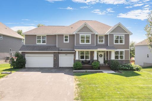 Sunlit front exterior highlights the home's well-maintained landscaping, gray siding, stone accents, and architectural gables. The inviting front porch, oversized driveway, and three-car garage offer excellent functionality and impressive curb appeal