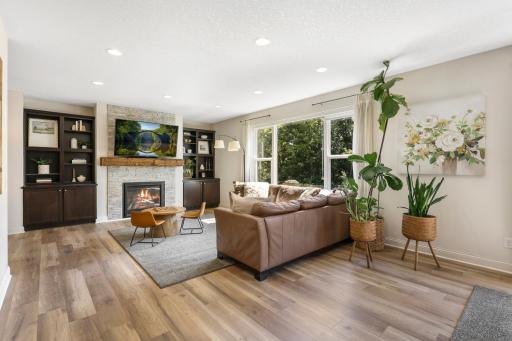 Warm and inviting living room featuring custom built-in cabinetry flanking a stacked-stone gas fireplace with a rustic wood mantel. Expansive floor-to-ceiling windows flood the space with natural light while offering lush backyard views