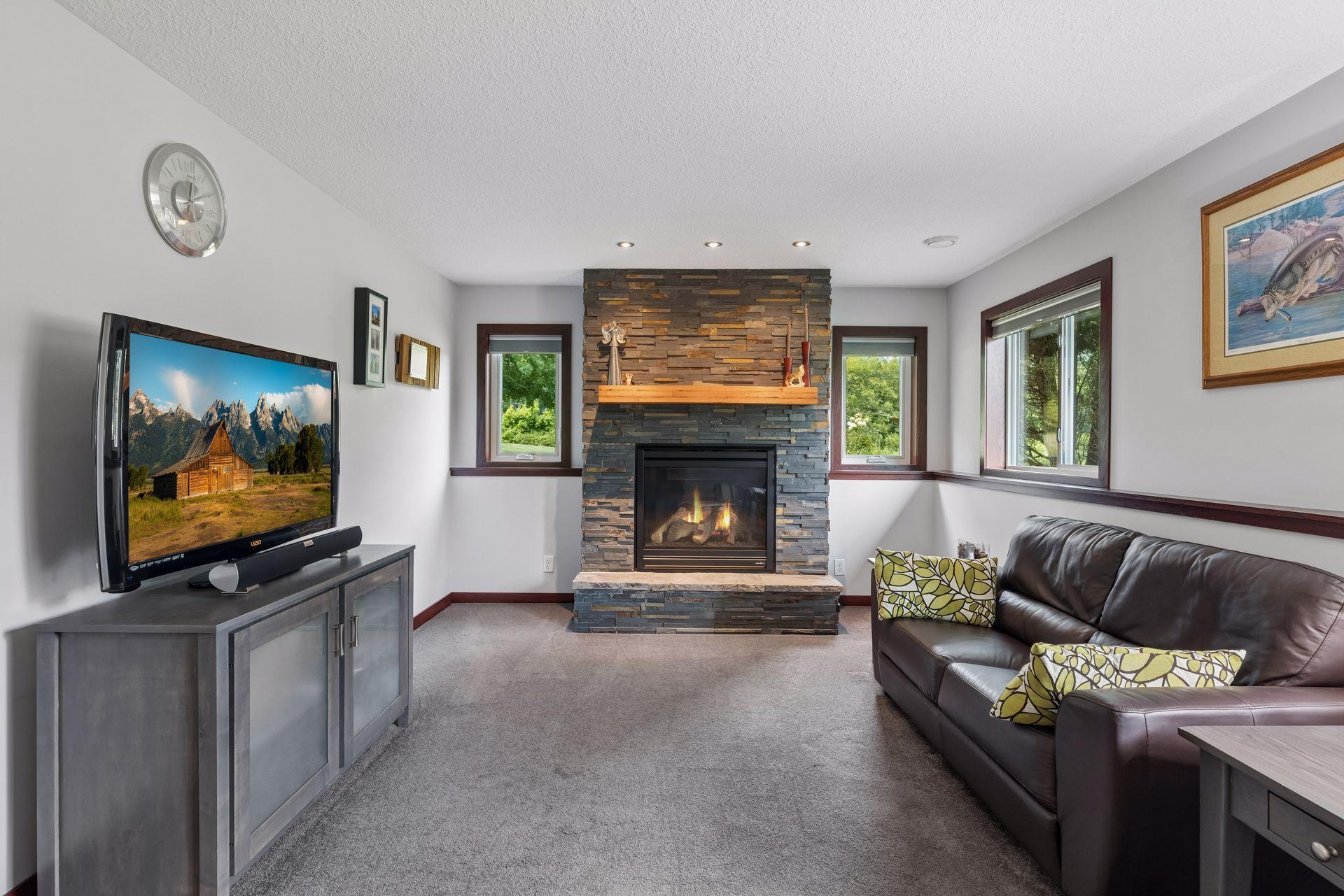 A gas fireplace with stone surround creates a welcoming focal point in the lower level family room, surrounded by four windows, carpet flooring, and recessed lighting.